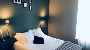 A neatly made bed with white and black bedding, two wall-mounted lamps, and framed pictures on a dark wall. - Maison Arquier (Aix-en-Provence)