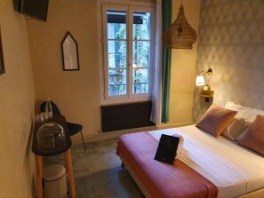 A hotel room with a bed, a window, a wall-mounted lamp, and a small table with a chair.