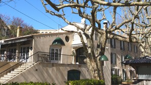 A building with a staircase leading to a balcony, a large tree in the foreground, and a clear blue sky. - Maison Arquier (Aix-en-Provence)