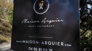 A black sign with the Maison Arquier hotel and restaurant logo, website, and contact number.