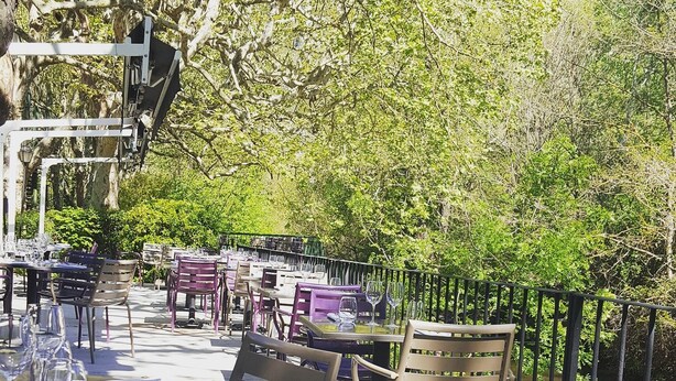 An outdoor dining area with tables and chairs, surrounded by greenery and a river. - Maison Arquier (Aix-en-Provence)