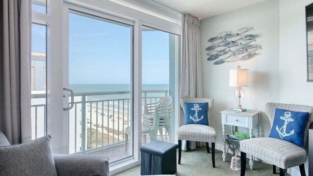A room with a large sliding glass door leading to a balcony, two upholstered chairs with anchor-themed pillows, a small side table with a lamp, and a wall-mounted fish decoration. - HS-1031 Experience ocean views, a spacious 2-BR condo in Myrtle Beach (Myrtle Beach)