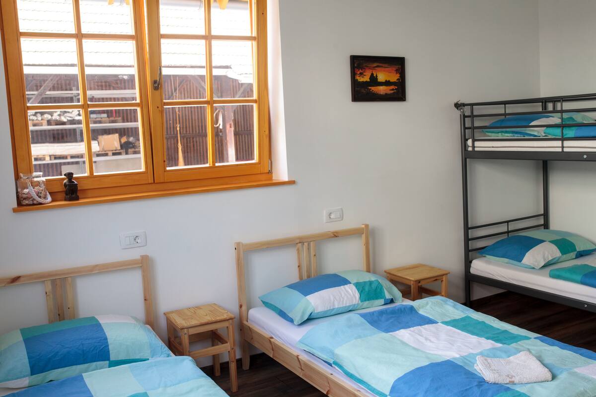 A room with two single beds, a bunk bed, a window with a view, a small table, and a wall-mounted light switch.