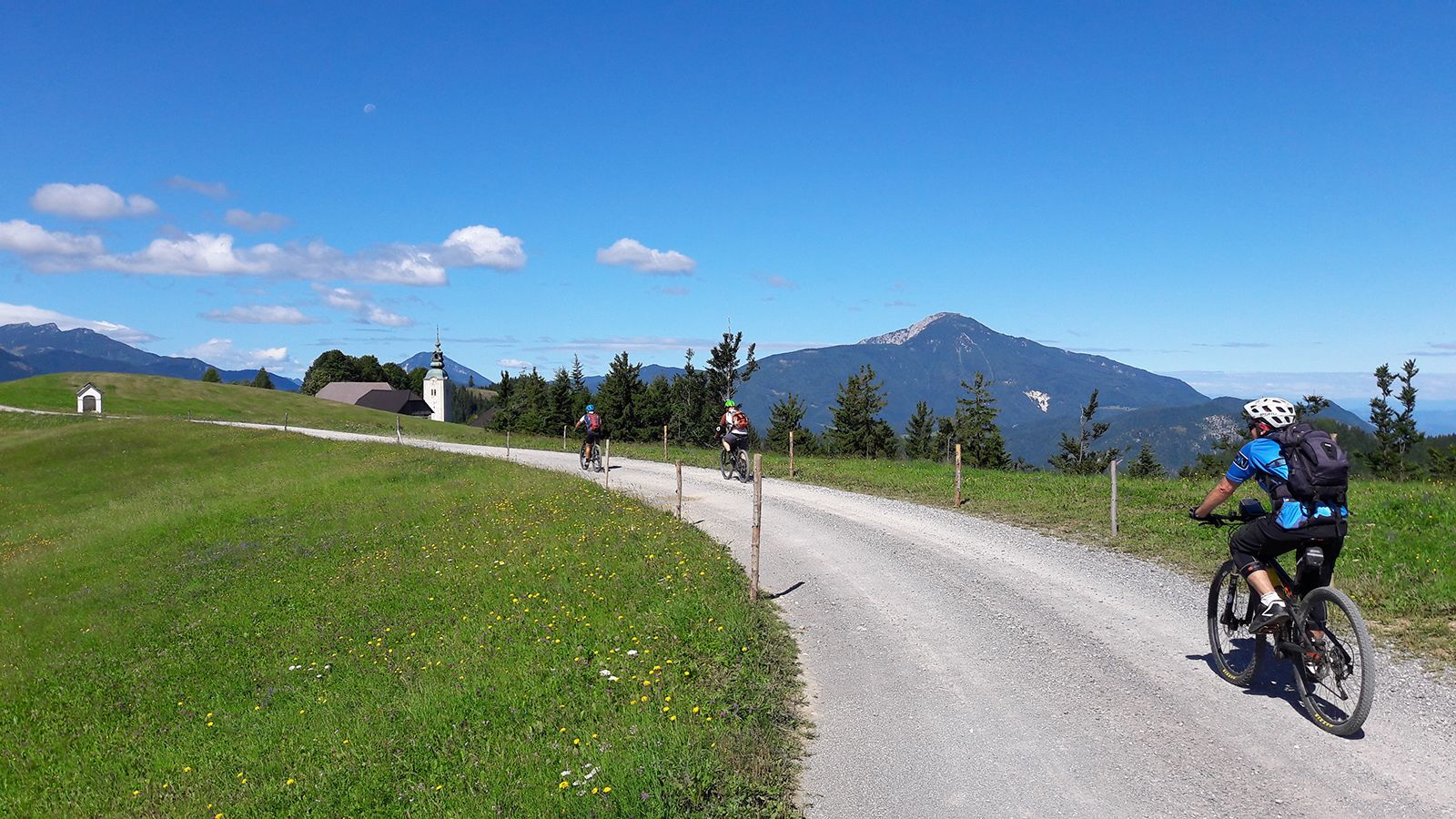 Un sendero de grava con ciclistas, rodeado de campos verdes y montañas al fondo.
