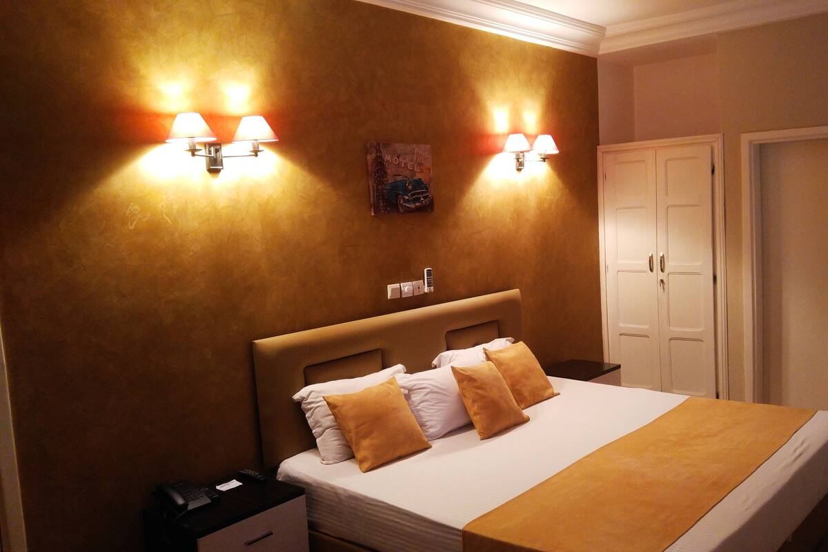 A hotel room with a large bed, two bedside lamps, a nightstand, and a wardrobe.