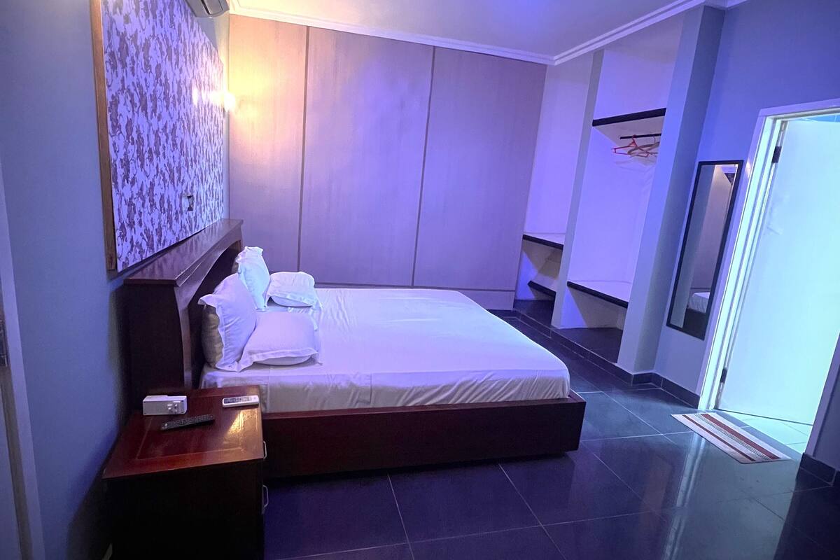 A modern hotel room with a large bed, a bedside table, a wall-mounted air conditioner, and a ceiling light.