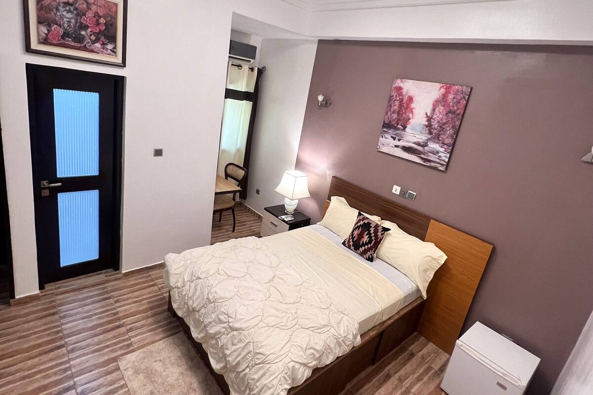 A bedroom with a bed, a nightstand, a lamp, a small white cabinet, a door with a window, and a painting on the wall.