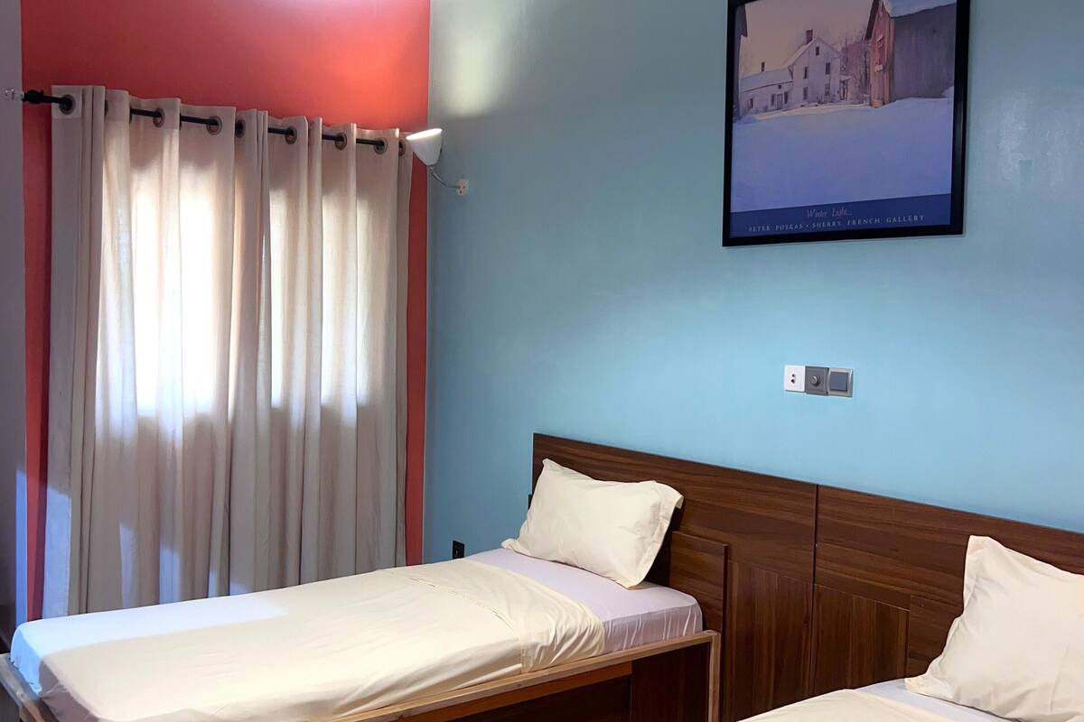 A hotel room with two beds, a ceiling light, a painting on the wall, and a window with curtains.