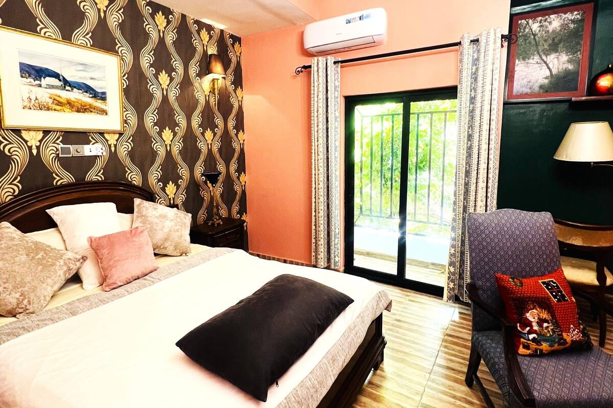 A hotel room with a bed, a chair, an air conditioning unit, and a view of greenery outside.