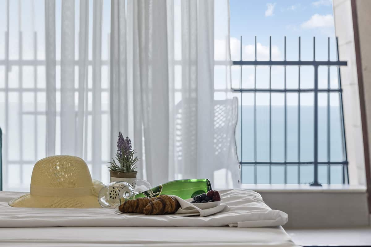 A bed with a straw hat, croissant, and a glass of juice, a small potted plant, and a green bottle on a white bedspread.