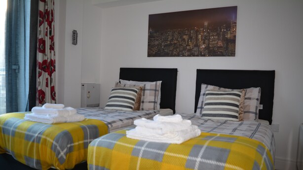A hotel room with two beds, each with a yellow and gray checkered bedspread and white towels. There is a cityscape painting on the wall above the beds. - Executive Apartment Cardiff Central (Cardiff)
