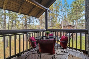 A wooden deck with a table and chairs, surrounded by a railing, overlooking a forested area.