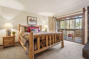 A bedroom with a wooden bed, a nightstand with a lamp, a window with a view of trees, and a balcony with outdoor furniture.