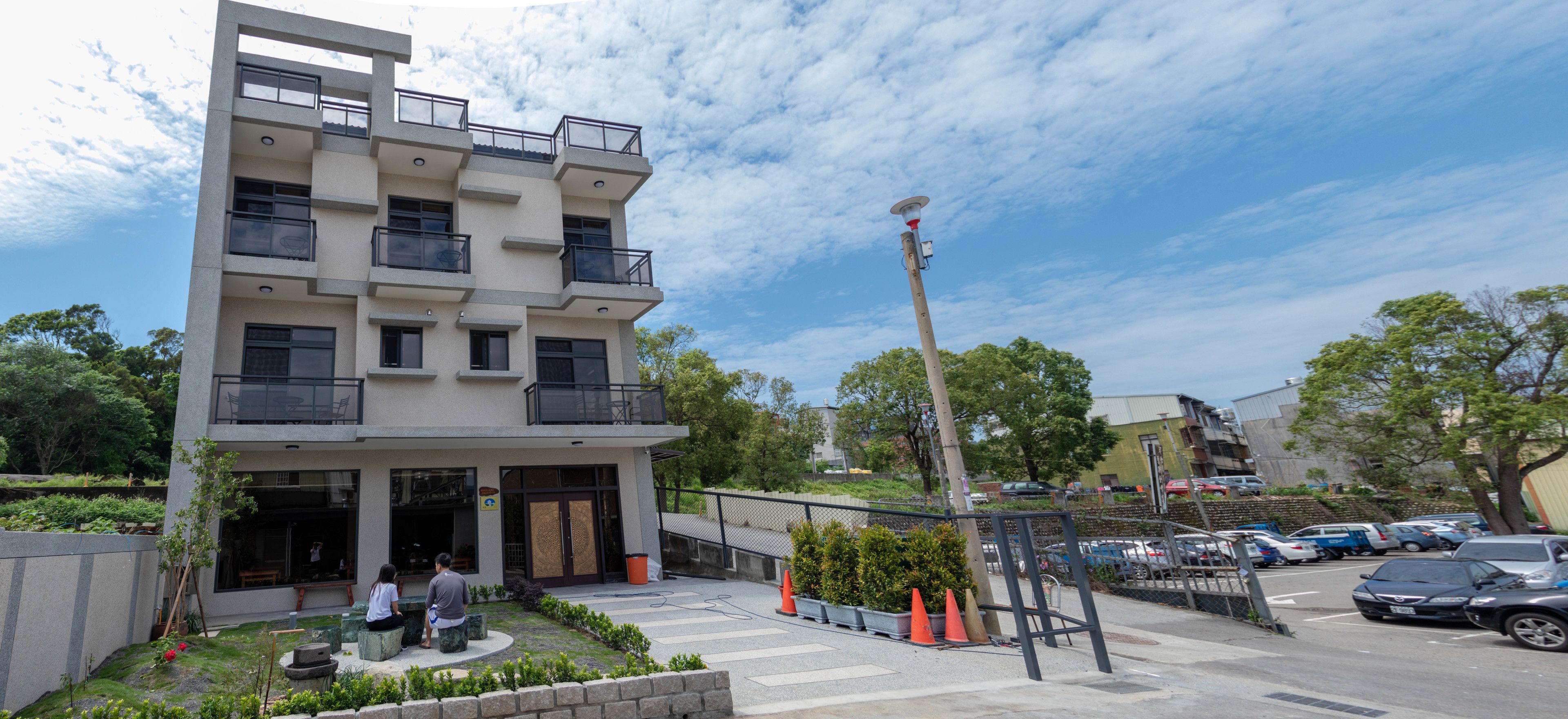 A modern multi-story building with balconies, a landscaped garden, and a parking area with cars.