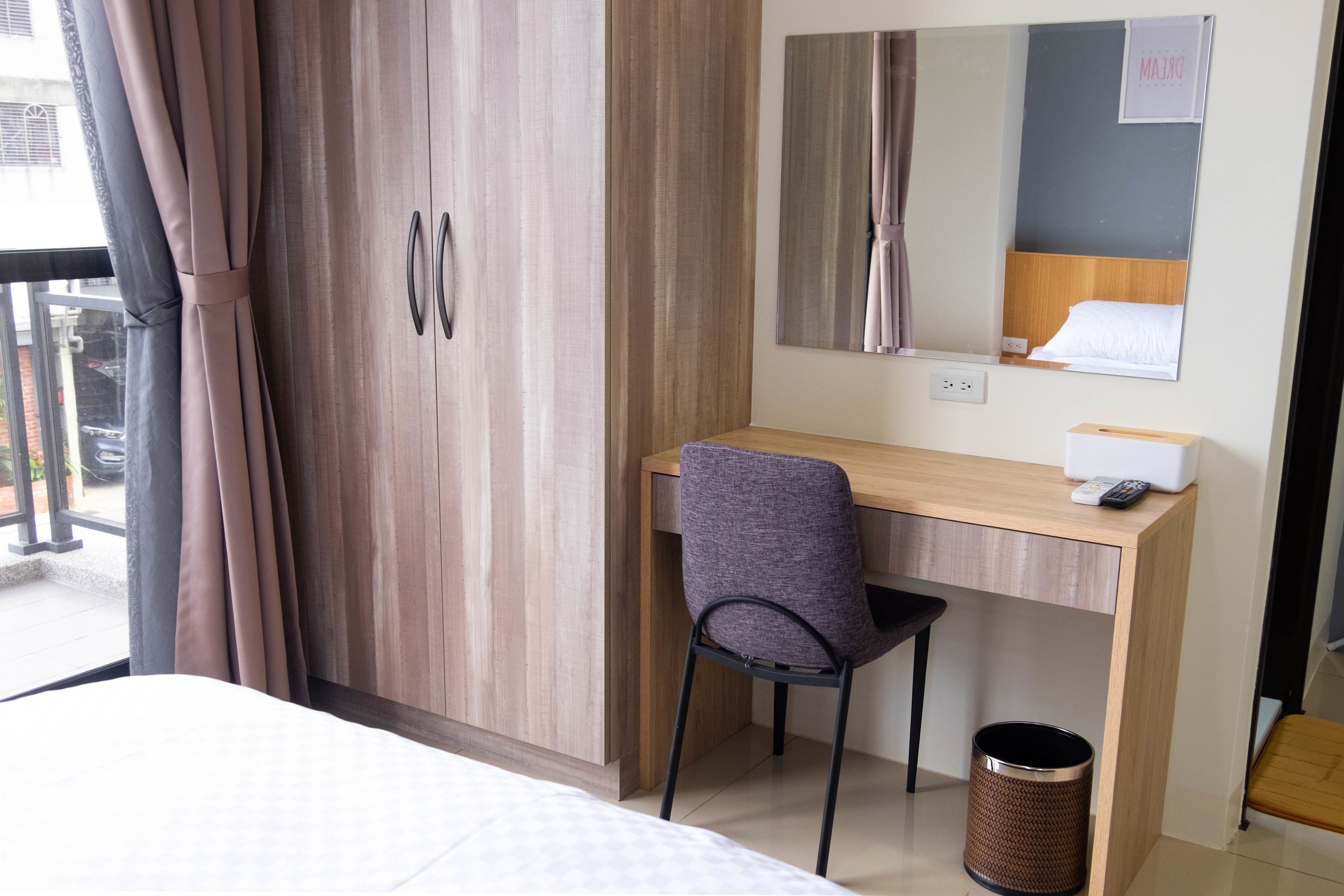 A hotel room with a wooden desk, a chair, a bed, a wardrobe, a mirror, and a trash bin.