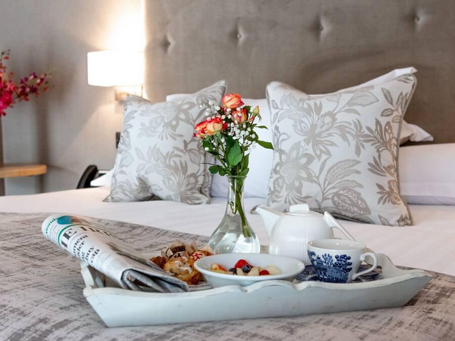 A neatly made breakfast tray with a newspaper, a vase of flowers, and a teapot on a bed with floral pillows.