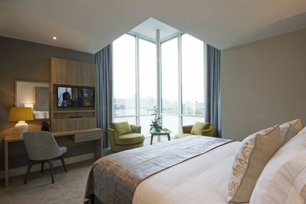 A hotel room with a large bed, a desk with a chair, a television, and a view of the city through large windows. - Gleesons Townhouse (Dublin)