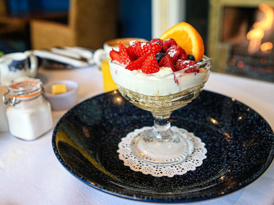 A dessert in a glass bowl on a black plate with a lace doily, surrounded by a table setting including a pitcher, a small bowl, and a jar.