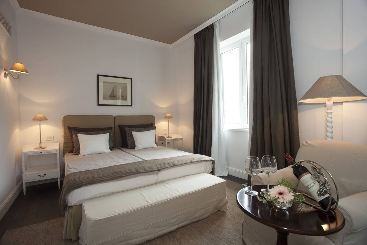 A hotel room with a bed, bedside tables with lamps, a chair, a small table with a wine bottle and glasses, and a window with curtains.