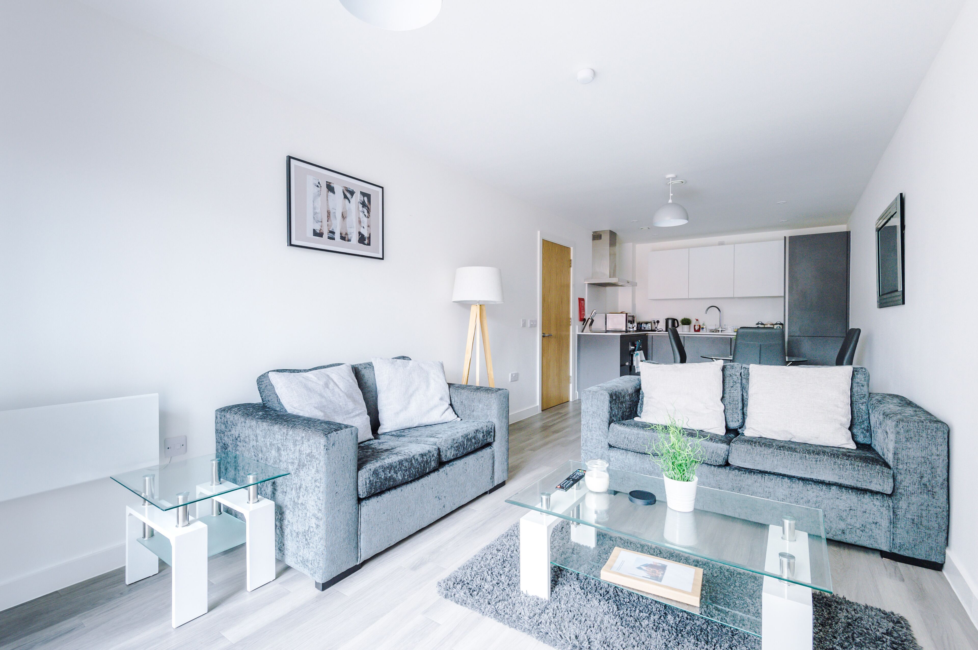 A modern living room with two grey sofas, a glass coffee table, and a kitchen in the background.