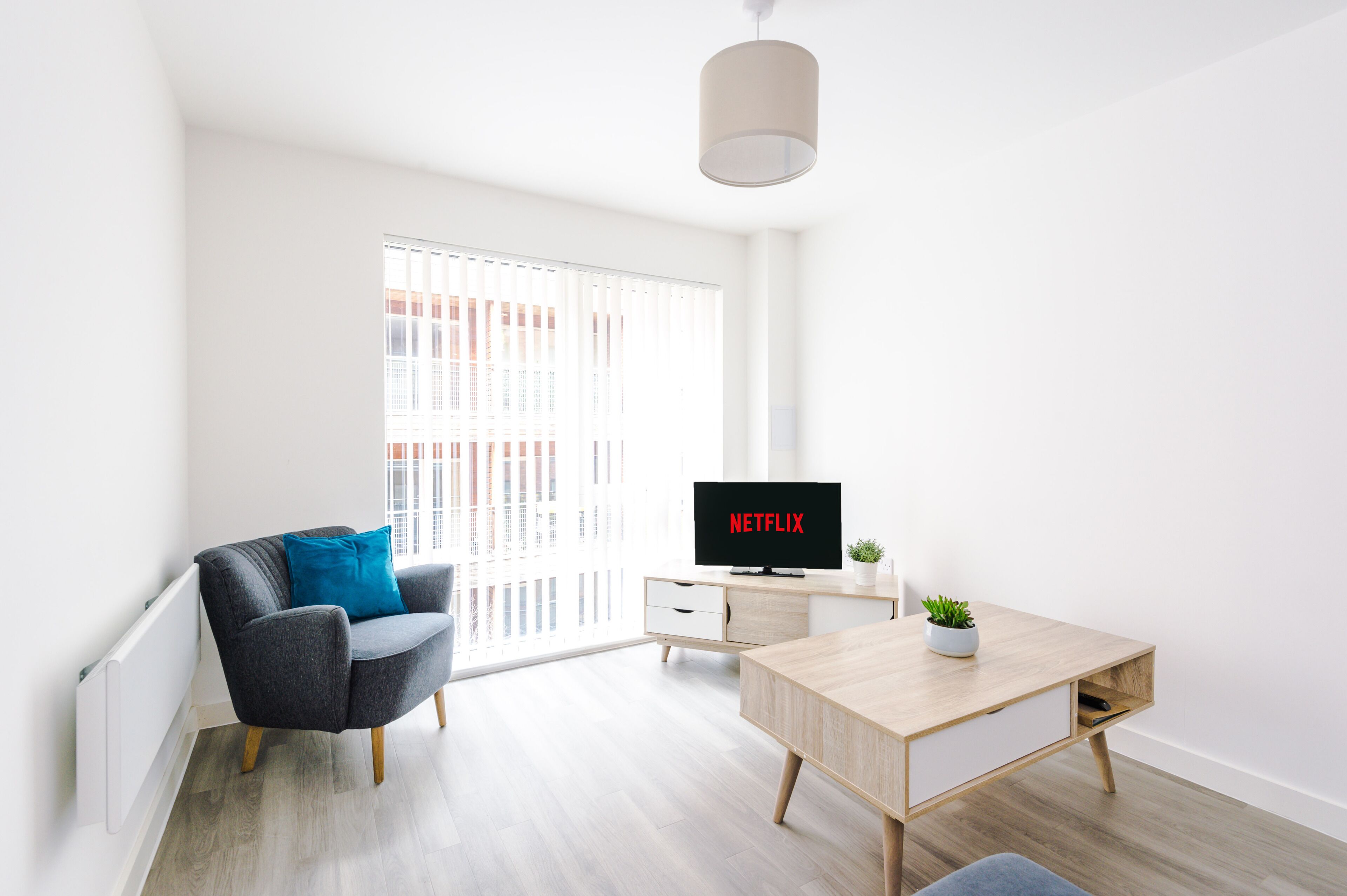 Un salon moderne avec un canapé gris, une table basse en bois, un téléviseur affichant le logo « NETFLIX » et une grande fenêtre avec des stores.