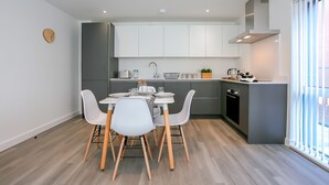 A modern kitchen with white cabinets, a dining table with chairs, and a large window with blinds. - Hilltop Serviced Apartments - Northern Quarter (Manchester)