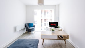 A modern living room with a sofa, a coffee table with a plant, a TV displaying 'NETFLIX', and a large window with a city view. - Hilltop Serviced Apartments - Northern Quarter (Manchester)