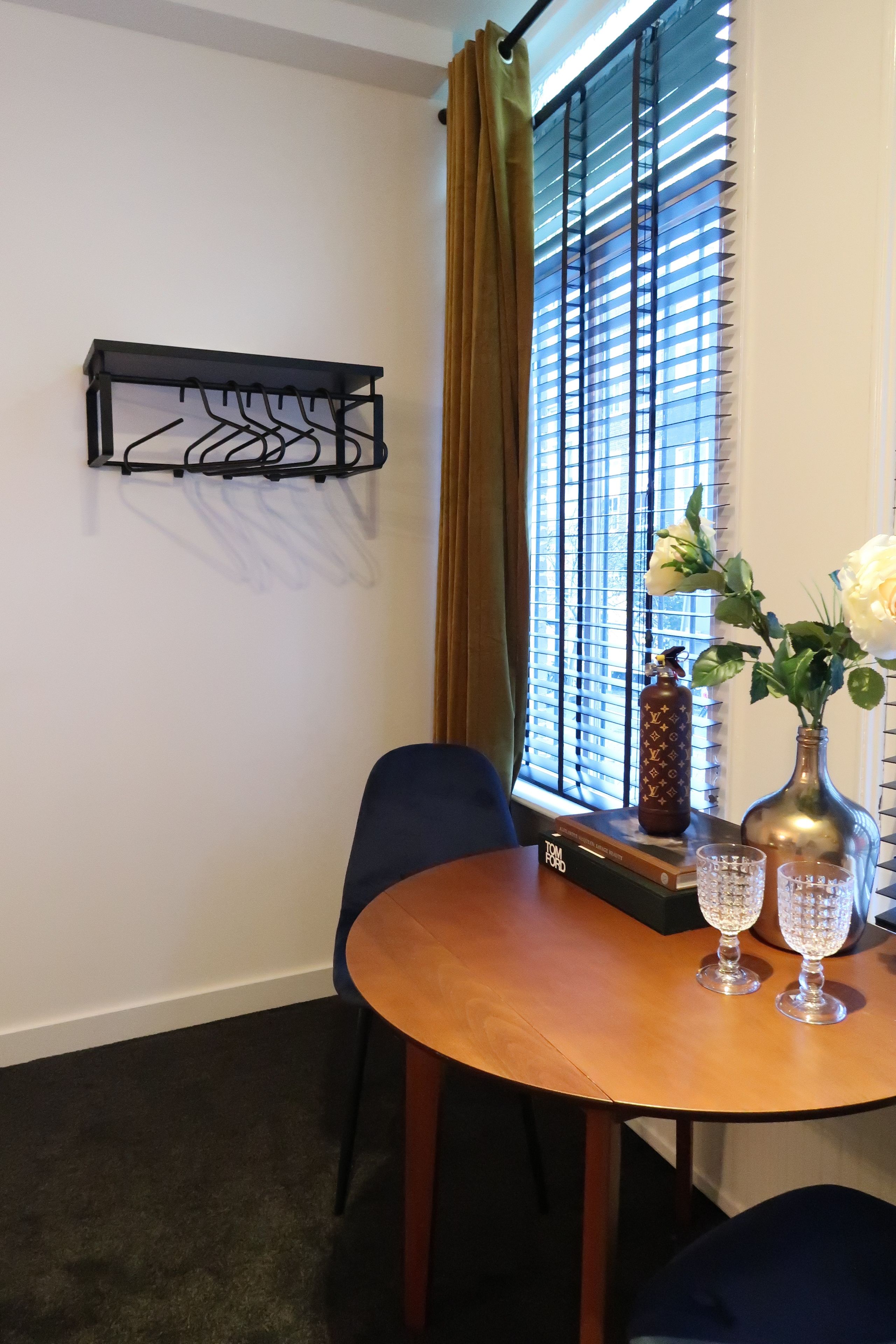 A dining area with a round wooden table, two chairs, a black coat rack, a vase with white flowers, and a Louis Vuitton bottle.