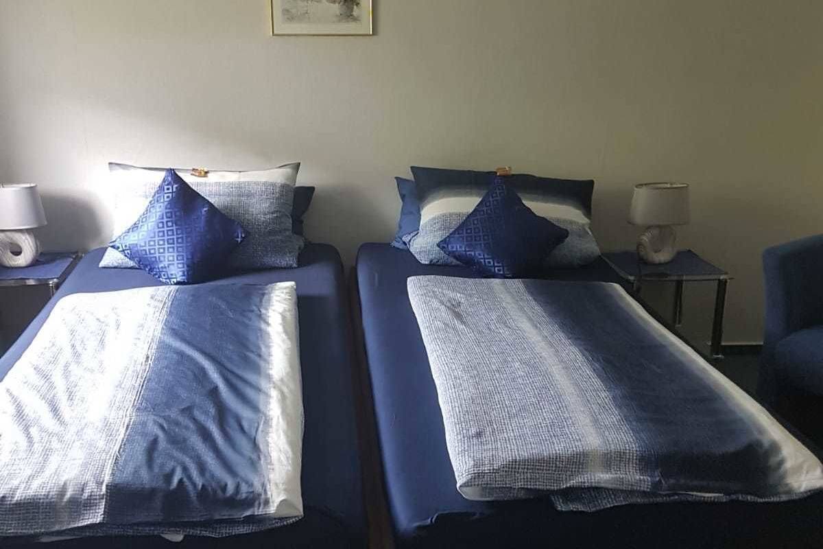 Una habitación con dos camas, cada una con ropa de cama y almohadas azules, una mesita con lámpara y un cuadro en la pared.