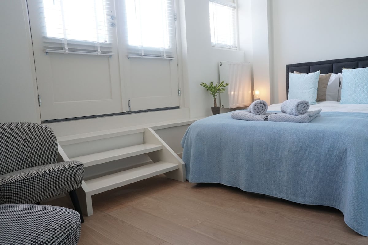 Un dormitorio con cama, una silla, una mesita de noche, una lámpara y una ventana.