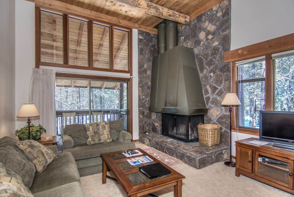 A cozy living room with a stone fireplace, wooden ceiling beams, and large windows.