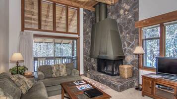 A cozy living room with a stone fireplace, wooden ceiling beams, and large windows.