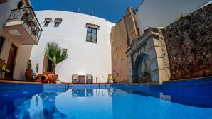 A swimming pool with a clear blue water surface, surrounded by a white building with a stone wall, a wooden door, and a small balcony with a metal railing. - Calergi 15: Boutique Resort 1BDR/1LVR Apt- Pool/Rooftop/Nature/Sunset/Rethymno (Ατσιπόπουλο)