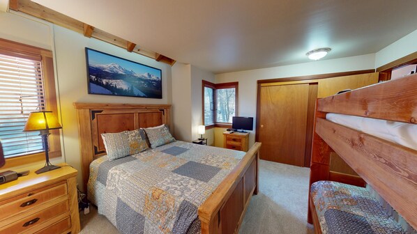 A bedroom with a wooden bed, a bedside table, a lamp, a TV, and a painting of a mountain landscape.