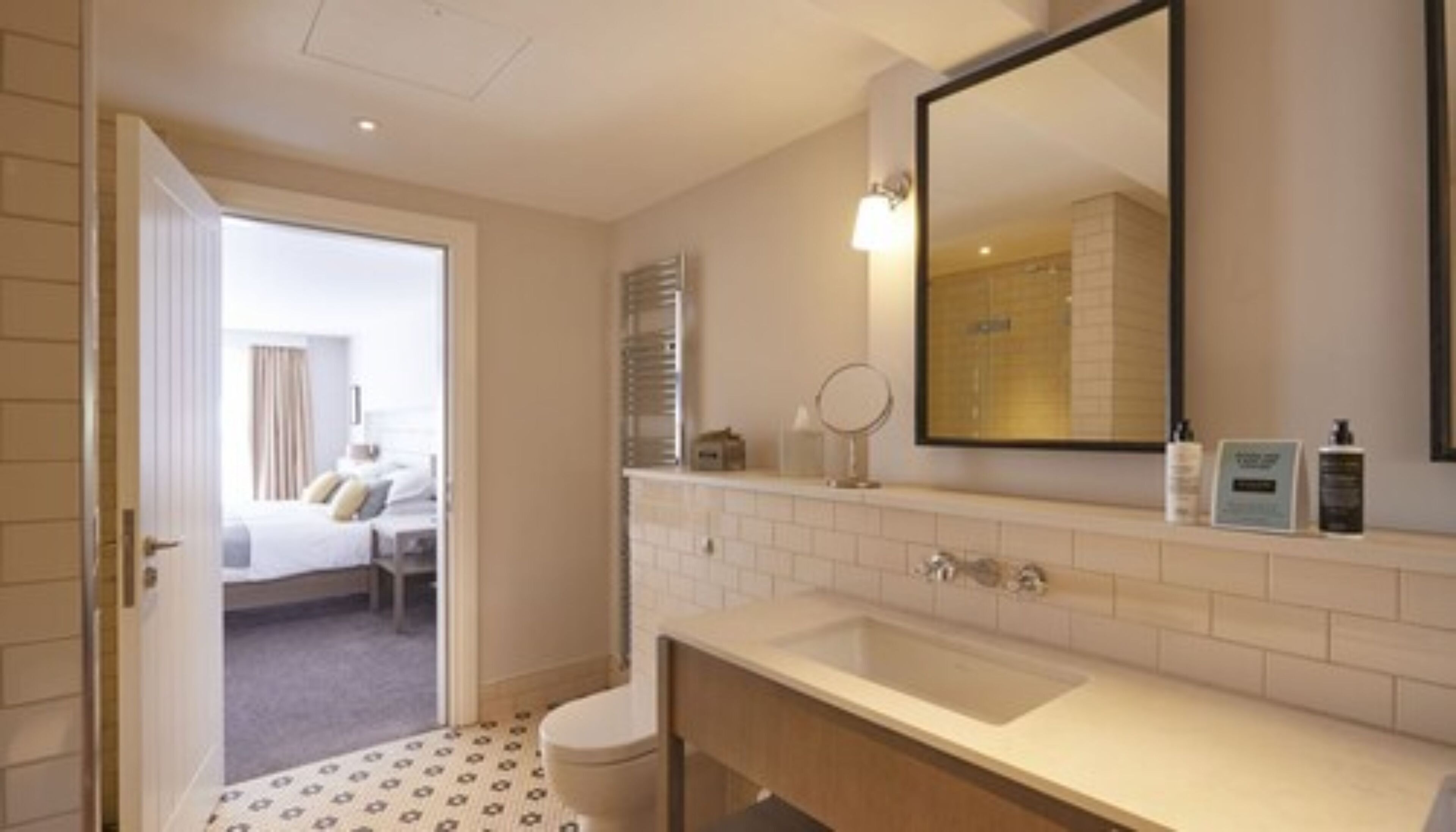 Un baño de hotel con un espejo grande, un lavamanos y un inodoro. Hay una puerta que da a un dormitorio con una cama y una ventana.