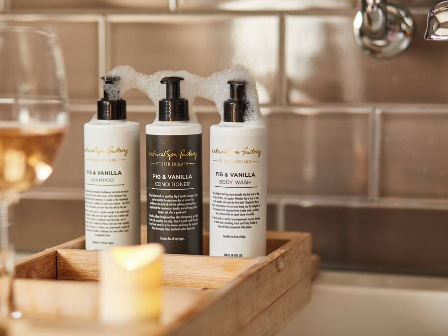Trois flacons de gel douche aux senteurs de figue et de vanille, un verre de vin et une bougie, le tout posé sur un plateau en bois.