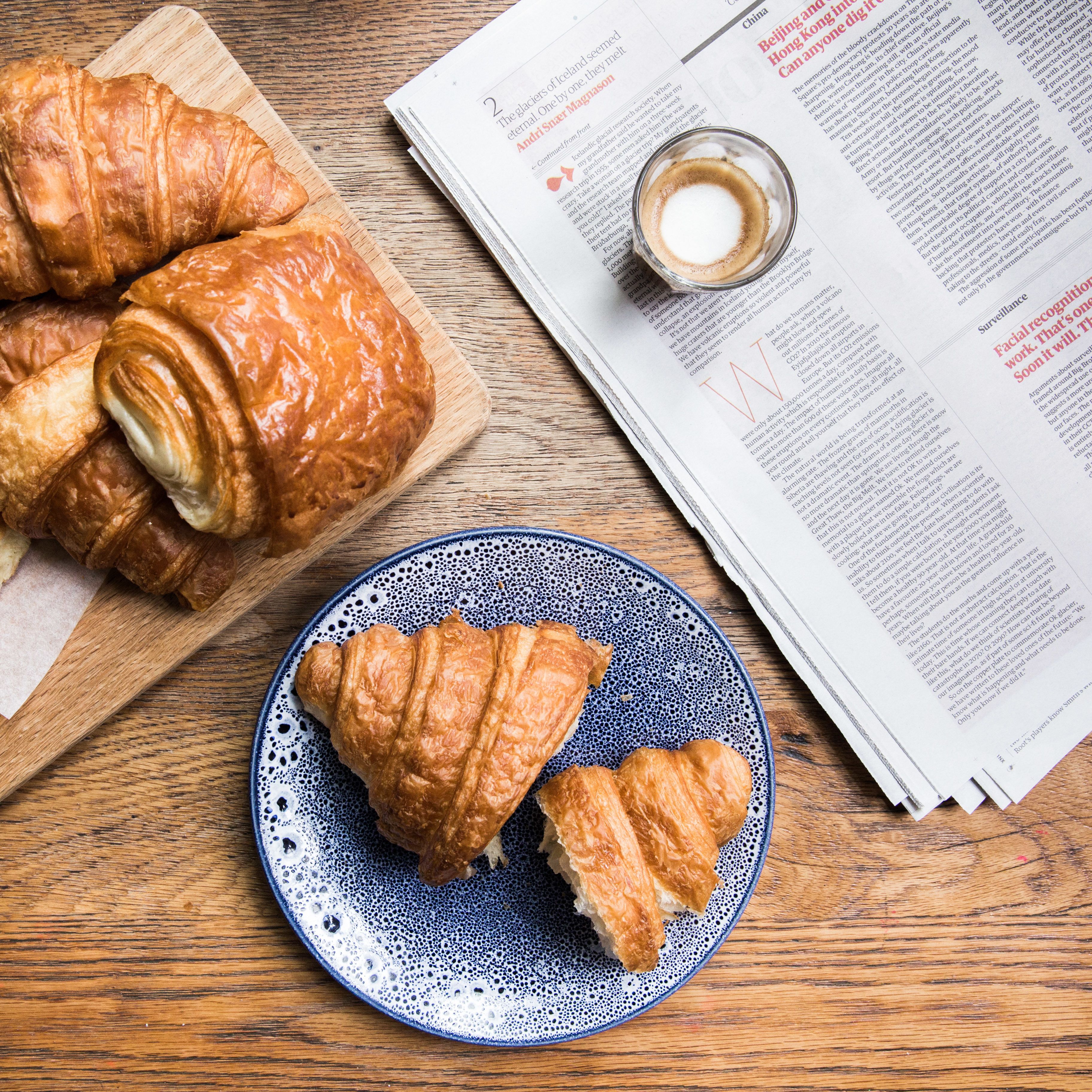 A wooden table with croissants, a plate with a croissant, a cup of coffee, and a newspaper.