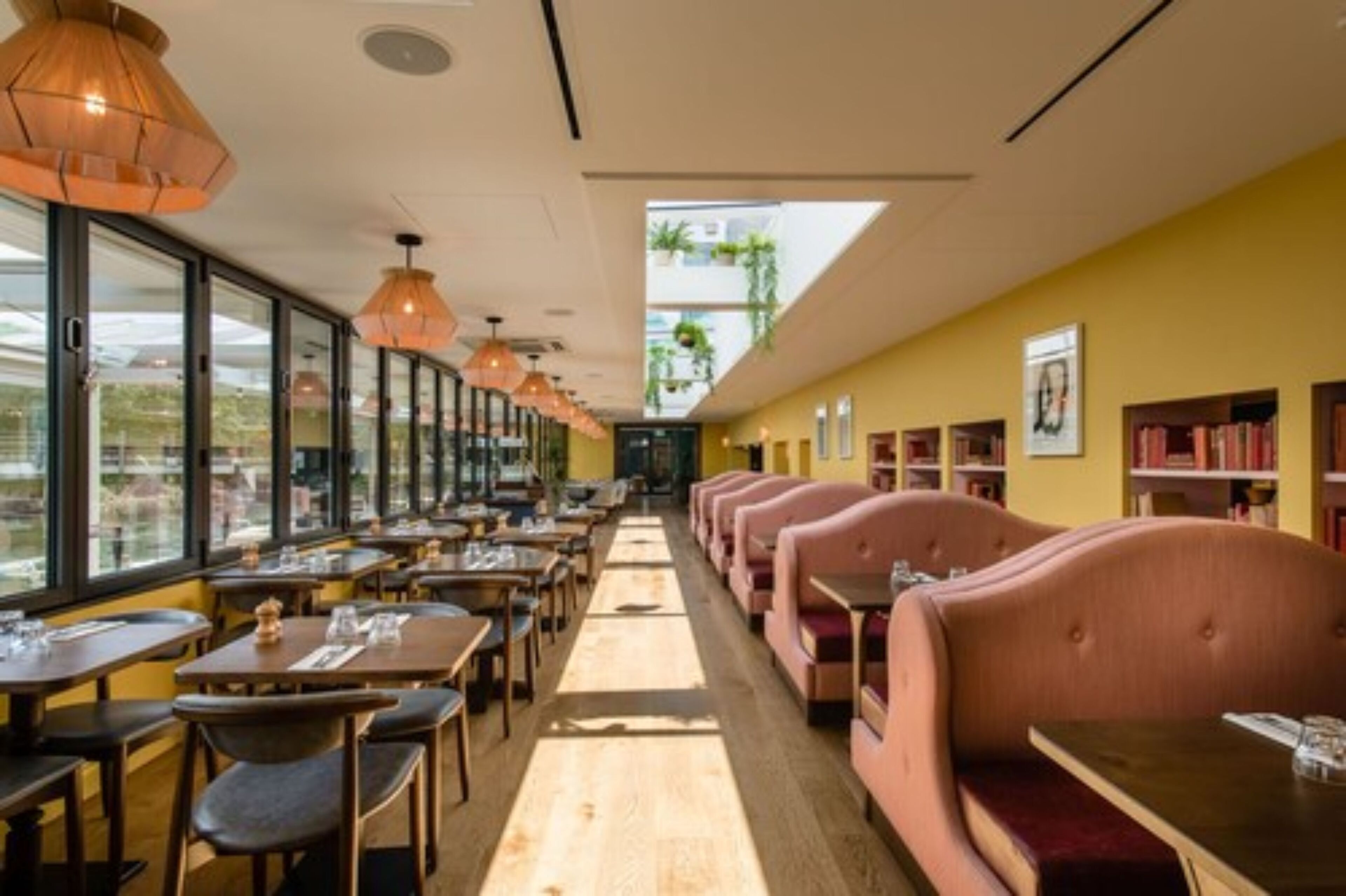 A restaurant with long tables and booths, large windows, and hanging pendant lights.