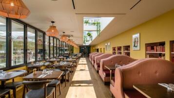A restaurant with long tables and booths, large windows, and hanging pendant lights.