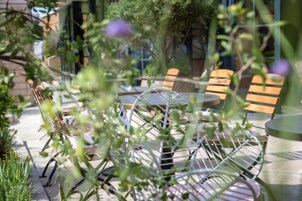 An outdoor seating area with metal chairs and tables, surrounded by greenery and a building in the background.