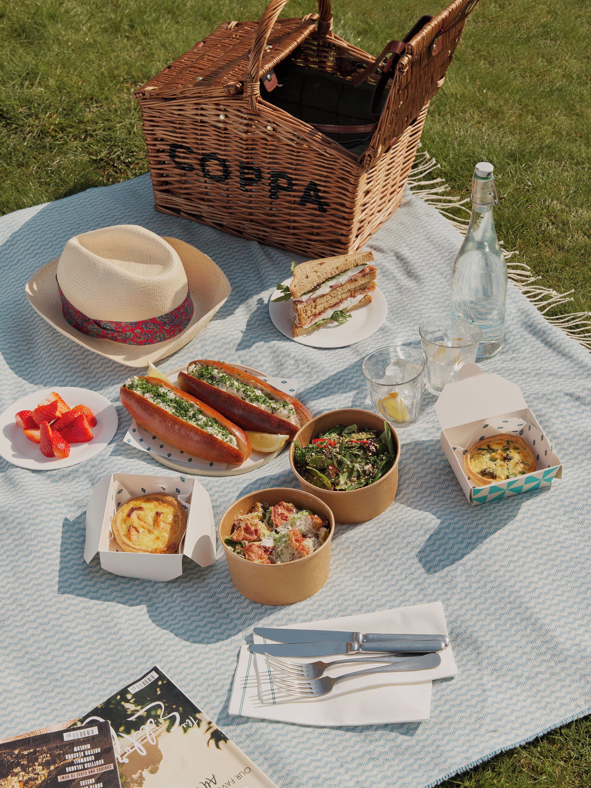 Un montaje para picnic con una cesta que dice "COPPA", un sombrero, sándwiches, perritos calientes, ensaladas y una botella, todo sobre una manta.