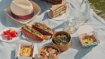 Un montaje para picnic con una cesta que dice "COPPA", un sombrero, sándwiches, perritos calientes, ensaladas y una botella, todo sobre una manta.