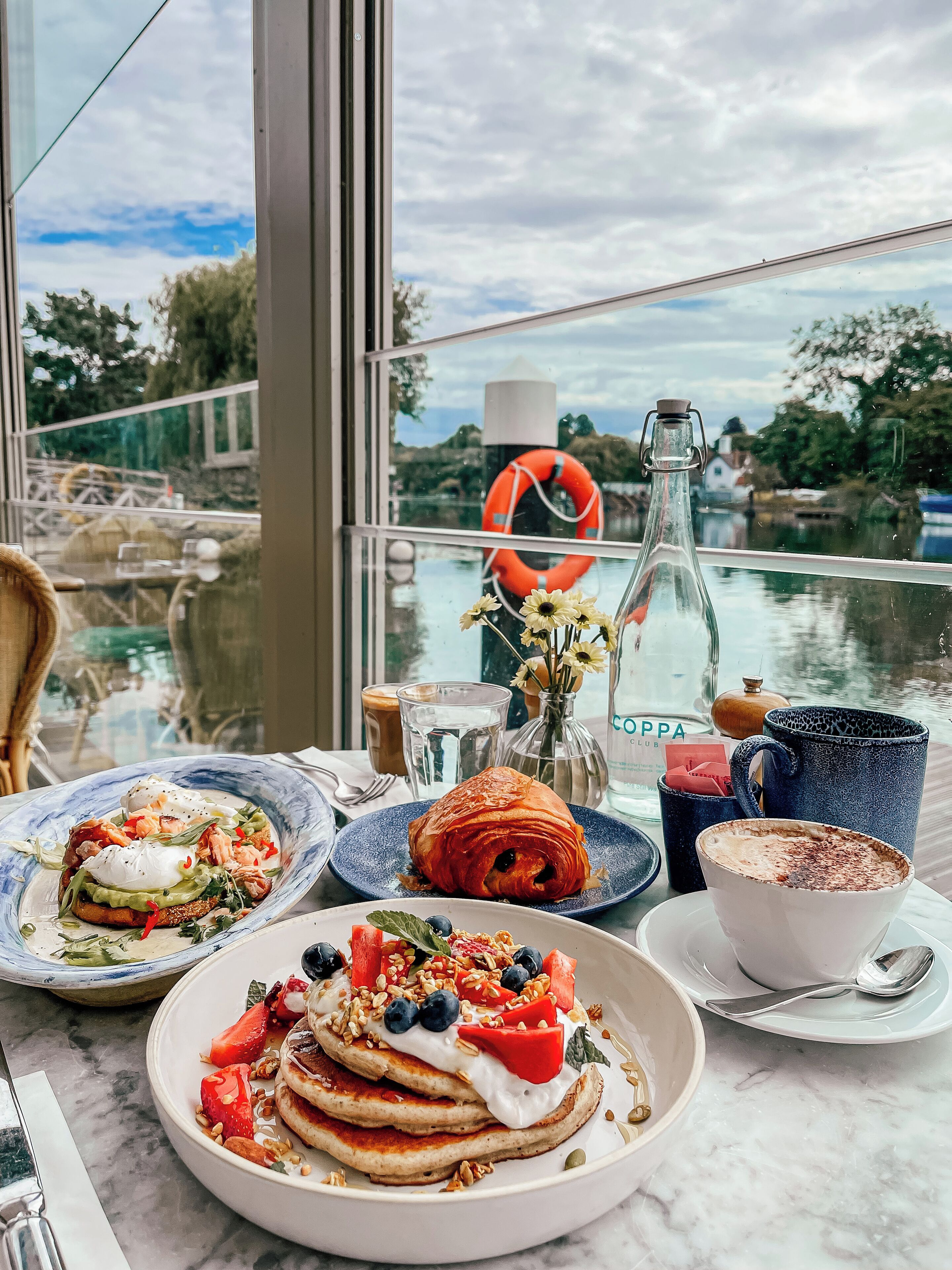 A table with breakfast items, a bottle, and a cup, with a view of a body of water and greenery outside.
