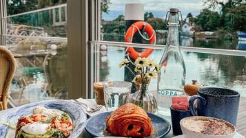 A table with breakfast items, a bottle, and a cup, with a view of a body of water and greenery outside.