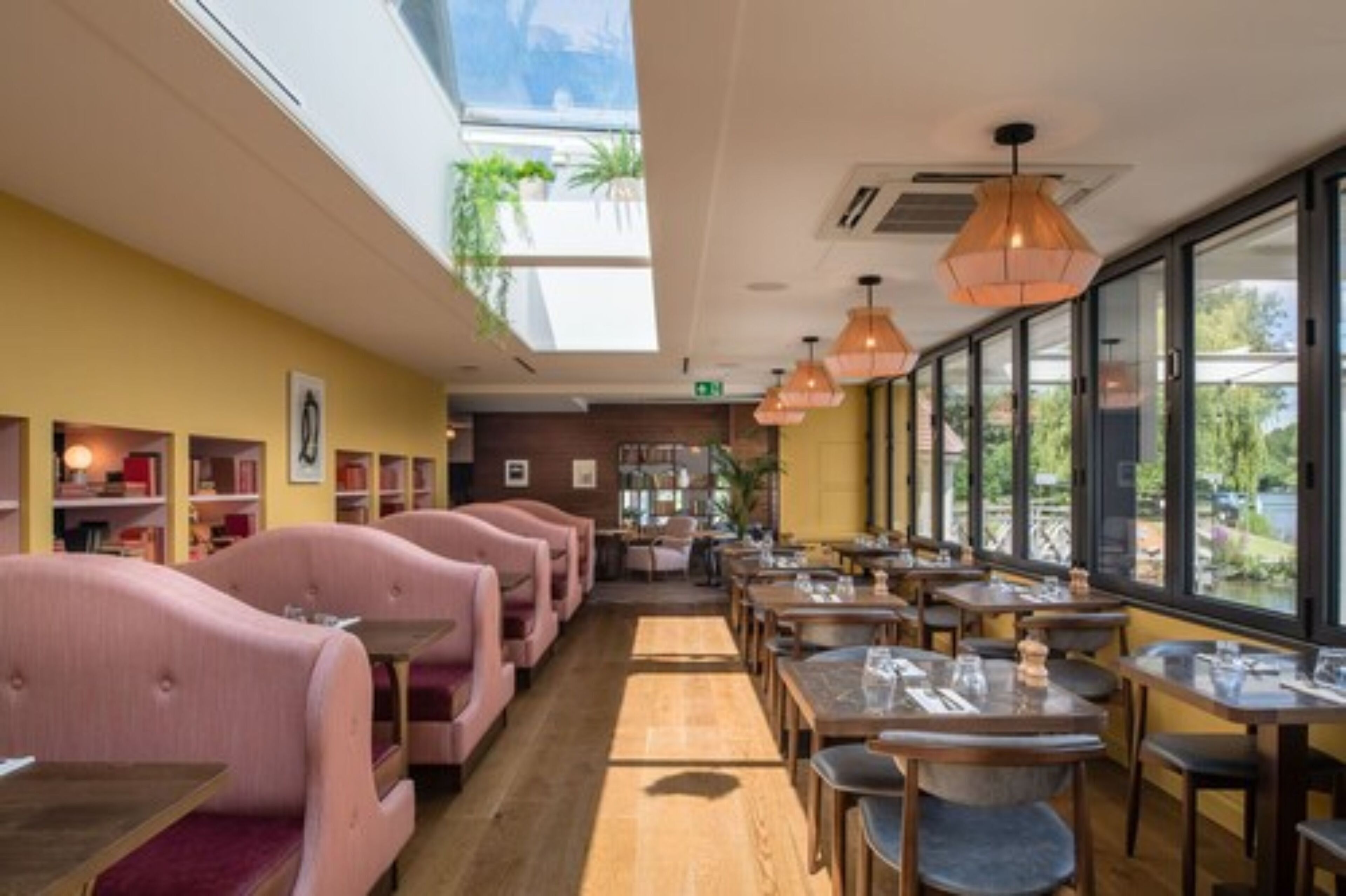 Un restaurante con asientos tapizados en rosa, mesas de madera, ventanas amplias y lámparas colgantes.