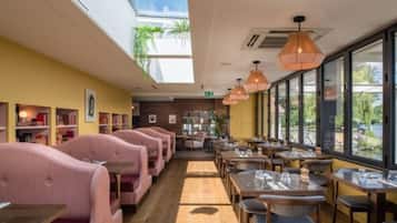 Un restaurante con asientos tapizados en rosa, mesas de madera, ventanas amplias y lámparas colgantes.