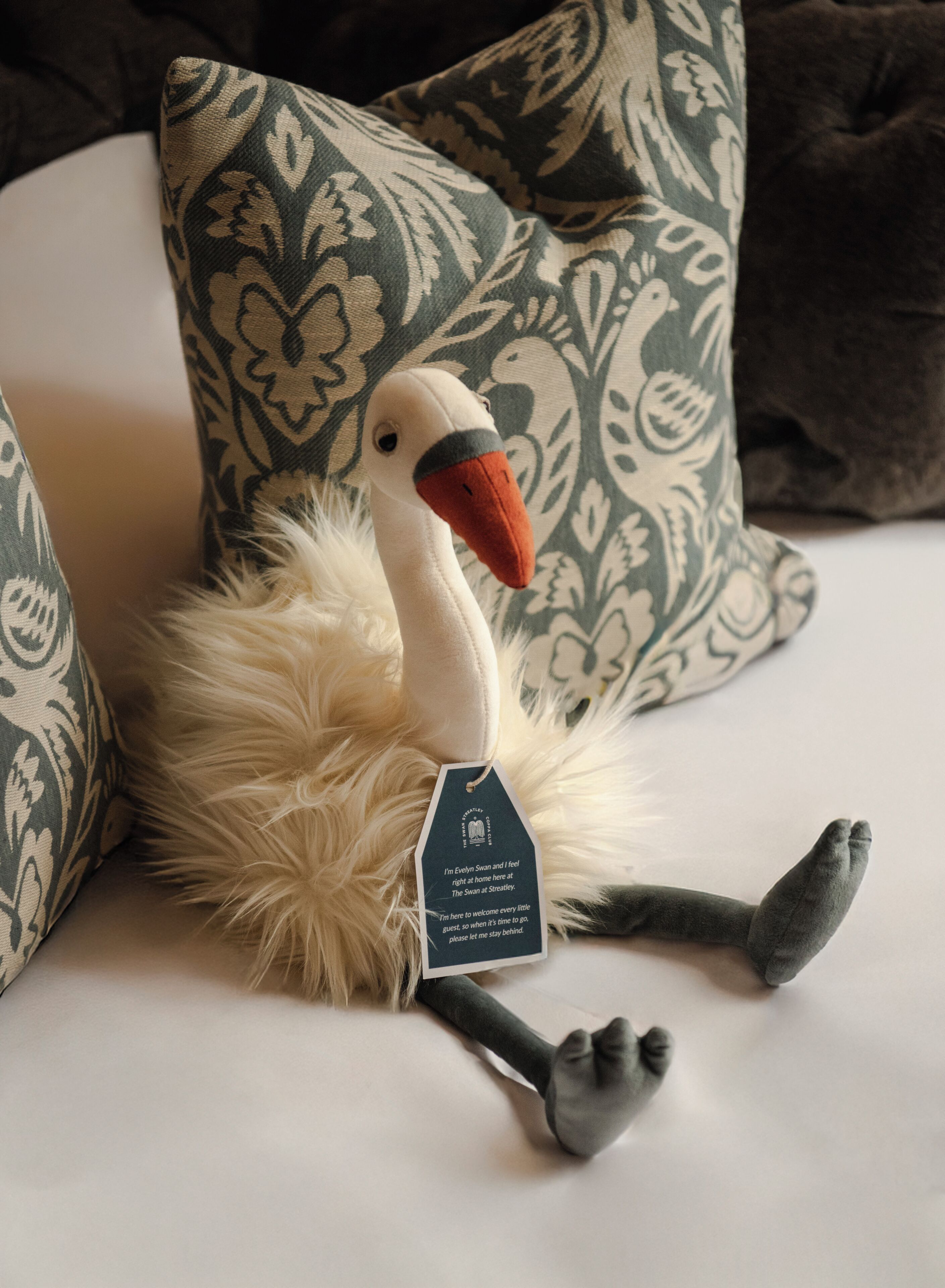 Un juguete de cisne suave con una etiqueta sobre una superficie blanca, rodeado de almohadones estampados.