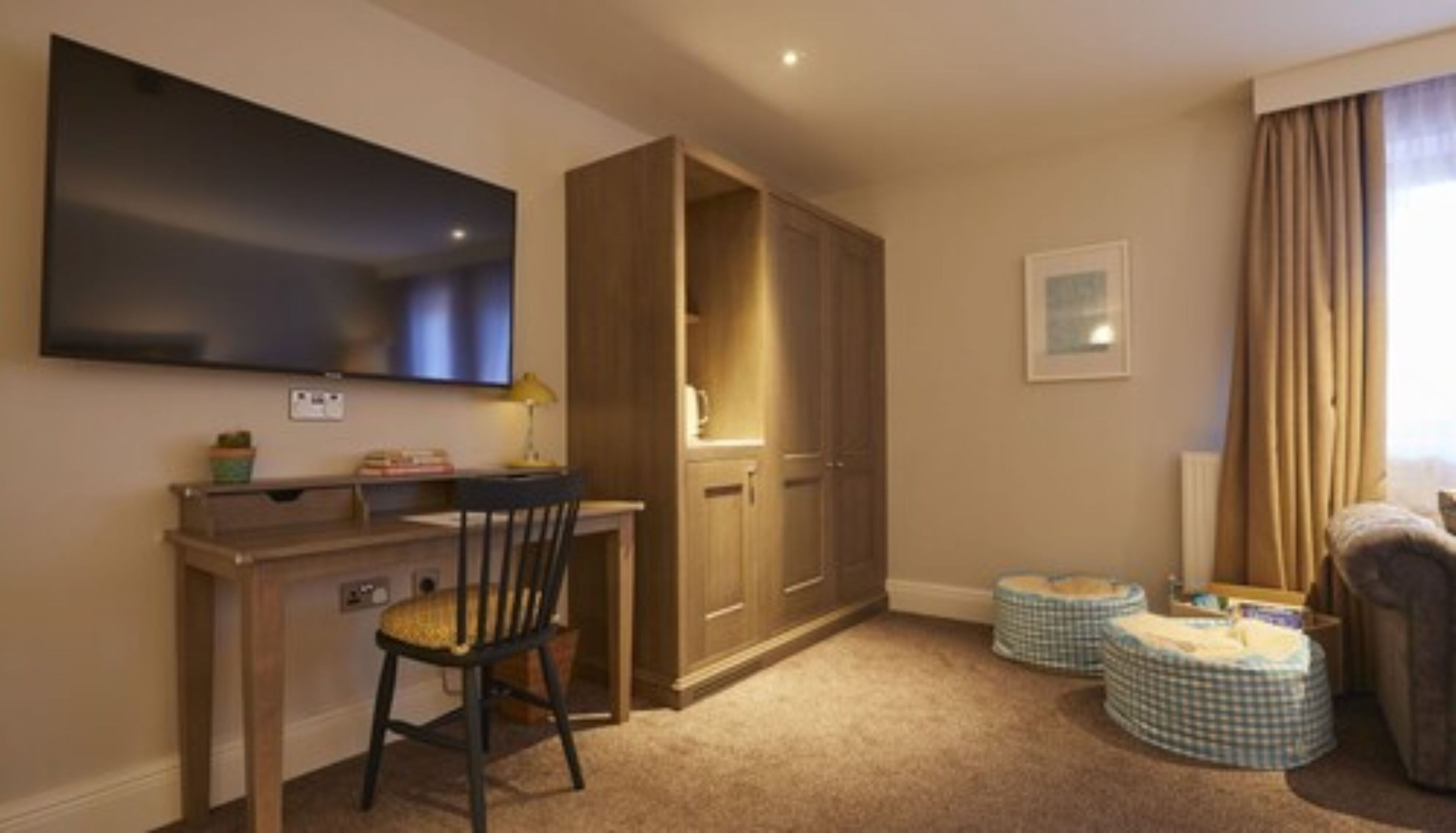 A hotel room with a flat-screen TV mounted on the wall, a wooden desk with a chair, a wardrobe, two checkered ottomans, and a window with curtains.