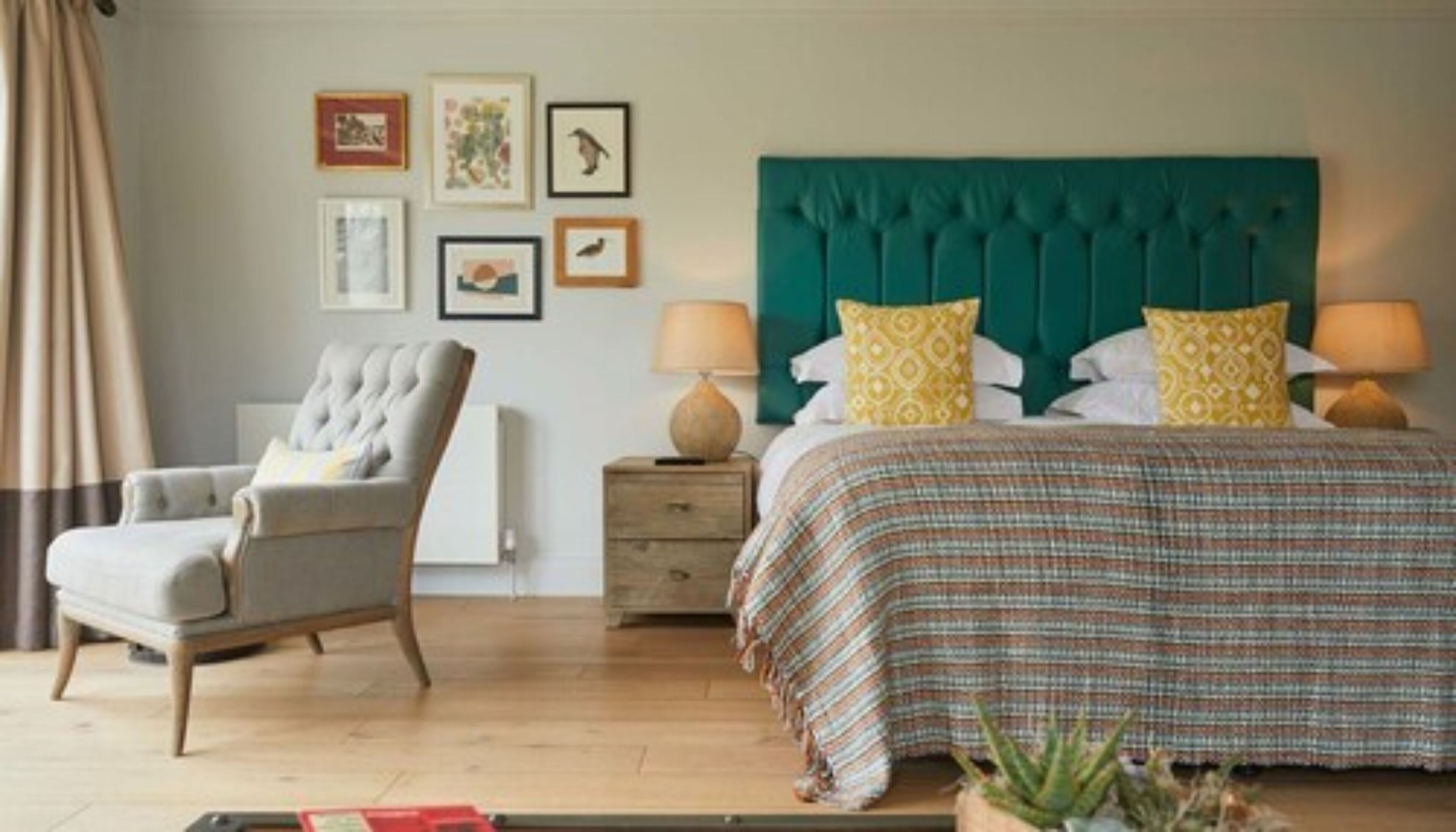 A bedroom with a tufted headboard, a bed with patterned bedding, a bedside table with lamps, a chair, and framed artwork on the wall.