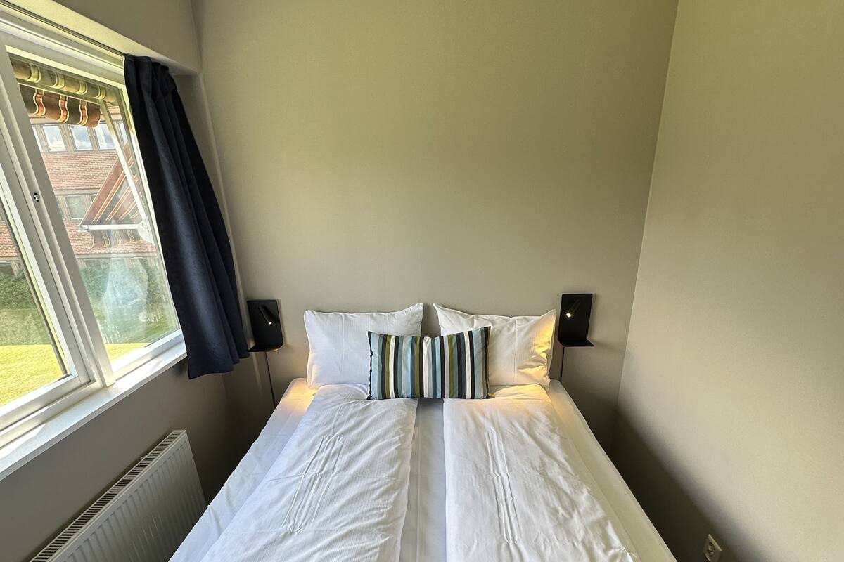 Una cama bien tendida con sábanas blancas y un plaid azul oscuro, una ventana con vistas a un edificio y un radiador.